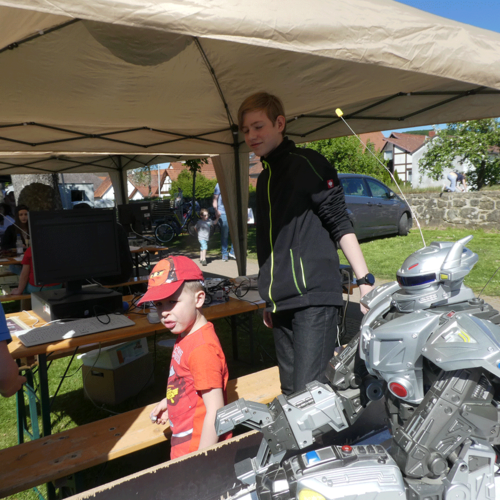 Kind und Jugendlicher an einer Bierzeltgarnitur unter einem Pavillion, im Vordergrund ist ein Spielzeug-Roborter zu sehen